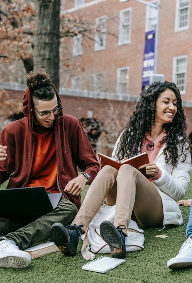 Drei Studierende sitzen auf einer Wiese, lesen Bücher und arbeiten an Laptops. Sie tragen lässige Kleidung und lächeln.