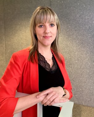 A woman with blonde hair is wearing a red blazer and stands with her arms crossed in front of a gray wall.