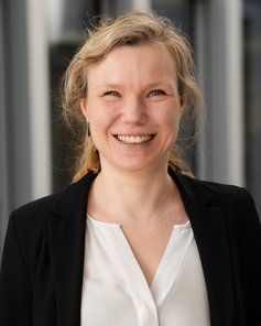 Eine lächelnde Frau mit blonden, lockigen Haaren trägt ein schwarzes Jackett über einem weißen Oberteil.