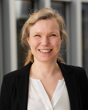 Eine lächelnde Frau mit blonden, lockigen Haaren trägt ein schwarzes Jackett über einem weißen Oberteil.