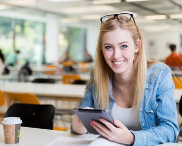 Eine blonde Frau mit Brille sitzt an einem Tisch, lächelt und nutzt ein Tablet. Eine Kaffeetasse steht neben ihr.