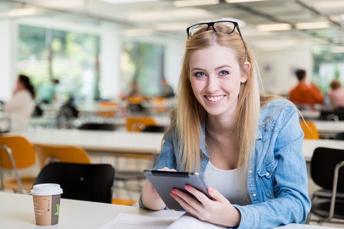Eine blonde Frau mit Brille sitzt an einem Tisch, lächelt und nutzt ein Tablet. Eine Kaffeetasse steht neben ihr.