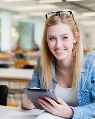 Eine blonde Frau mit Brille sitzt an einem Tisch, lächelt und nutzt ein Tablet. Eine Kaffeetasse steht neben ihr.