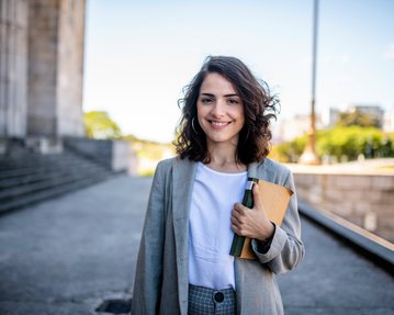 Eine junge Frau mit lockigem Haar trägt einen Blazer und hält ein Notizbuch sowie einen Stift in der Hand, während sie lächelt.