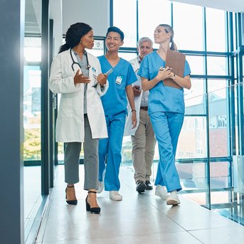 Eine Gruppe von vier medizinischen Fachkräften in blauer Arbeitskleidung geht durch einen modernen Flur im Krankenhaus.