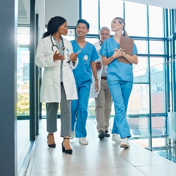 Eine Gruppe von vier medizinischen Fachkräften in blauer Arbeitskleidung geht durch einen modernen Flur im Krankenhaus.