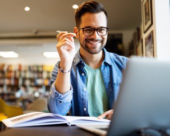 A man with glasses sits at a table, smiling and holding a pen while looking at a laptop and taking notes.