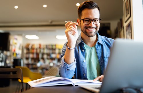 Ein junger Mann mit Brille sitzt an einem Tisch, lächelt und hält einen Stift in der Hand, während er auf einen Laptop schaut.
