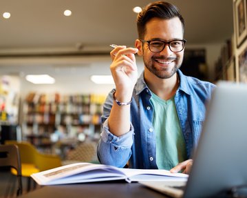 Ein junger Mann mit Brille sitzt an einem Tisch, lächelt und hält einen Stift in der Hand, während er auf einen Laptop schaut.