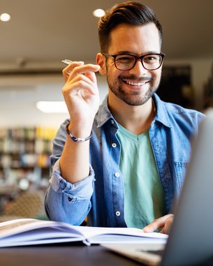 A man with glasses sits at a table, smiling and holding a pen while looking at a laptop and taking notes.