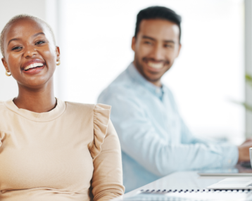 A smiling woman with short hair is sitting at a table while a man in the background is working on a laptop.
