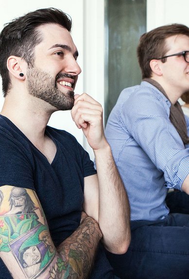 Eine Gruppe von fünf Personen sitzt an Tischen in einem Klassenzimmer. Ein Mann mit einem Bart und einem bunten Tattoo lächelt, während er aufmerksam zuhört. Die anderen Teilnehmer scheinen ebenfalls interessiert zu sein.