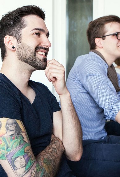 A group of five people is sitting at tables in a classroom. A man with a beard and a colorful tattoo is smiling while listening attentively. The other participants also seem to be engaged.