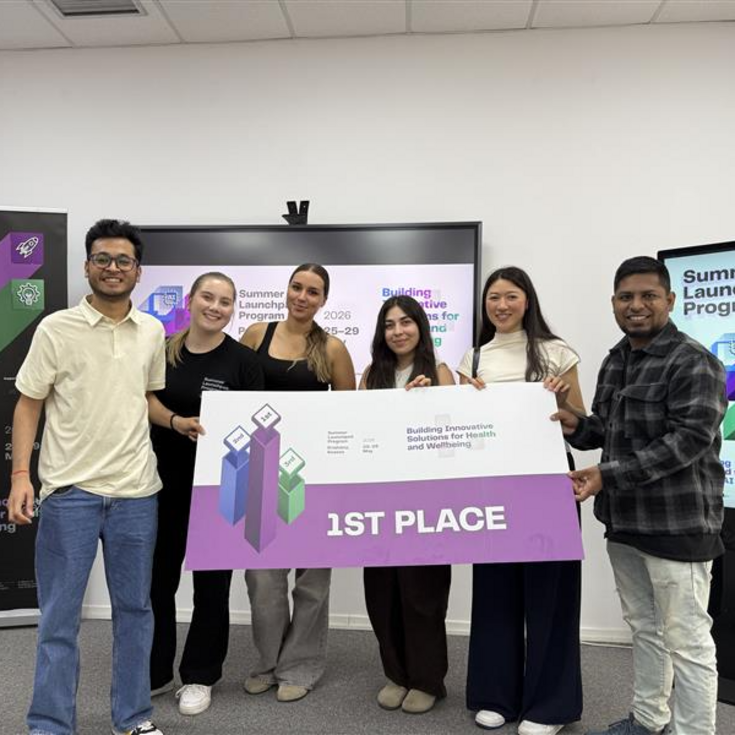 Sechs Personen stehen zusammen und halten ein großes Schild mit der Aufschrift "1ST PLACE" während einer Preisverleihung im Rahmen eines Programms.