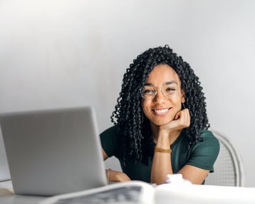 Kategorie Informatik SRH University Eine lächelnde Frau mit lockigem Haar sitzt an einem Tisch vor einem Laptop und schaut freundlich in die Kamera. Sie trägt eine grüne Bluse und eine Brille.