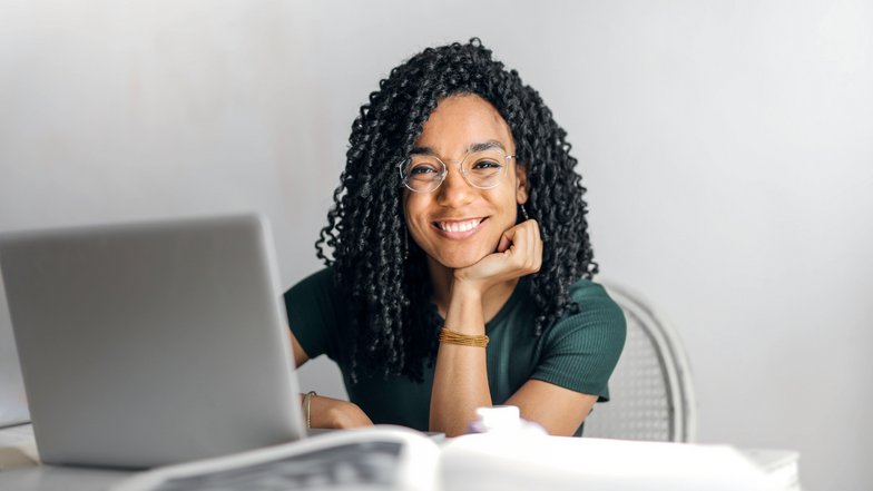Eine lächelnde Frau mit lockigem Haar sitzt an einem Tisch vor einem Laptop und schaut freundlich in die Kamera. Sie trägt eine grüne Bluse und eine Brille.