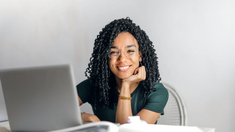 Eine lächelnde Frau mit lockigem Haar sitzt an einem Tisch vor einem Laptop und schaut freundlich in die Kamera. Sie trägt eine grüne Bluse und eine Brille.