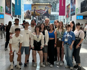 Gruppe von Personen posiert in einer großen Halle mit bunten Bannern und Menschen im Hintergrund.