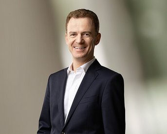 Ein Mann in einem dunklen Anzug mit weißem Hemd lächelt freundlich. Der Hintergrund ist unscharf und hell.