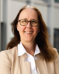 Eine Frau mit langen, braunen Haaren und Brille lächelt freundlich. Sie trägt ein helles Blazer und eine weiße Bluse.