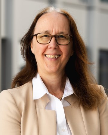 Eine Frau mit langen, braunen Haaren und Brille lächelt freundlich. Sie trägt ein helles Blazer und eine weiße Bluse.