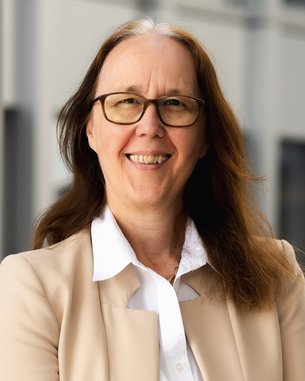 A woman with long brown hair and glasses smiles warmly. She is wearing a light blazer and a white blouse.