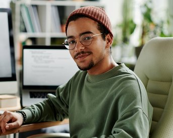 A young man with glasses and a cap sits at a desk, surrounded by computers and notes, looking into the camera.