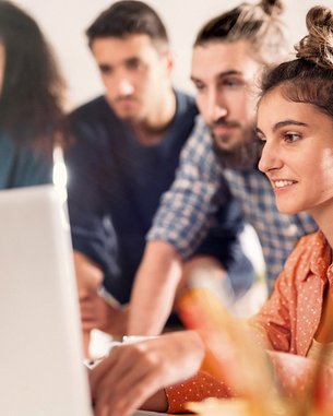 Eine Gruppe von vier Personen steht um einen Laptop. Eine Frau mit lockigem Haar und einem orangefarbenen Hemd arbeitet am Computer, während die anderen aufmerksam zuschauen. Die Umgebung wirkt modern und hell.