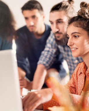 A group of four people is gathered around a laptop. A woman with curly hair and an orange shirt is working on the computer while the others watch attentively. The surroundings appear modern and bright.
