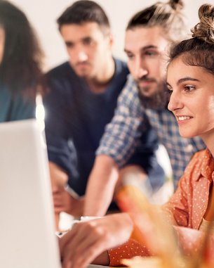 Eine Gruppe von vier Personen steht um einen Laptop. Eine Frau mit lockigem Haar und einem orangefarbenen Hemd arbeitet am Computer, während die anderen aufmerksam zuschauen. Die Umgebung wirkt modern und hell.