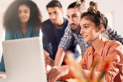 A group of four people is gathered around a laptop. A woman with curly hair and an orange shirt is working on the computer while the others watch attentively. The surroundings appear modern and bright.