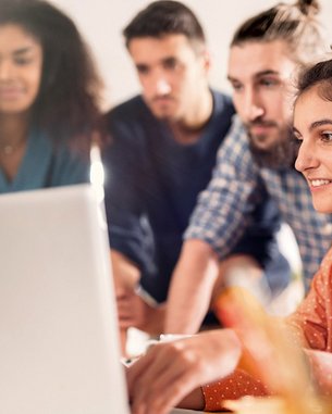 Eine Gruppe von vier Personen steht um einen Laptop. Eine Frau mit lockigem Haar und einem orangefarbenen Hemd arbeitet am Computer, während die anderen aufmerksam zuschauen. Die Umgebung wirkt modern und hell.