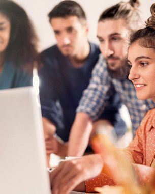 A group of four people is gathered around a laptop. A woman with curly hair and an orange shirt is working on the computer while the others watch attentively. The surroundings appear modern and bright.