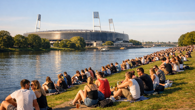 Menschen sitzen am Flussufer, genießen die Aussicht auf ein Stadion im Hintergrund. Es ist ein sonniger Tag mit vielen Besuchern.