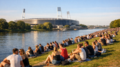 Menschen sitzen am Flussufer, genießen die Aussicht auf ein Stadion im Hintergrund. Es ist ein sonniger Tag mit vielen Besuchern.