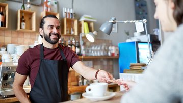 A barista hands a customer a cup of coffee at the counter of a café. The barista is wearing a black apron and smiling.
