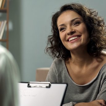 A smiling woman with curly hair is sitting across from another person, speaking while looking at a notepad.