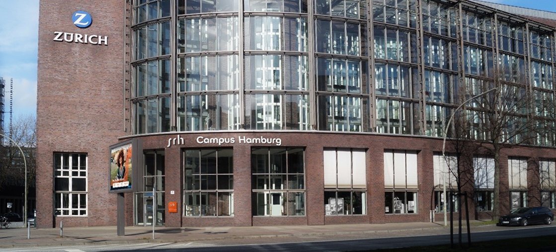 Moderne Glasfassade des SRH Campus Homburg mit dem Logo der Zurich Versicherung. Das Gebäude hat einen runden Turm und ist von einer hellen Umgebung umgeben.