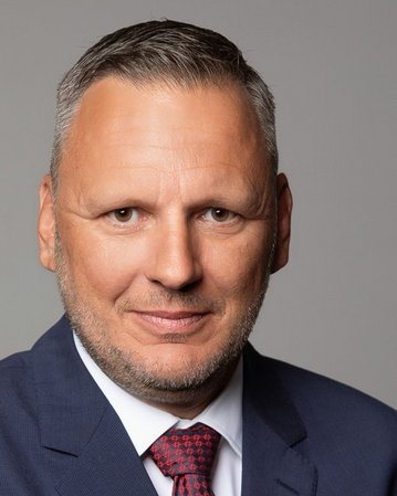 Ein Mann mit kurzen, grauen Haaren und einem gepflegten Bart trägt einen blauen Anzug und eine rote Krawatte.