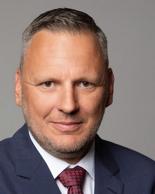 Ein Mann mit kurzen, grauen Haaren und einem gepflegten Bart trägt einen blauen Anzug und eine rote Krawatte.