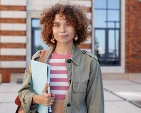Young woman with curly hair standing in front of a modern building, holding a binder in her hand. She is wearing a green jacket and has a shoulder bag.
