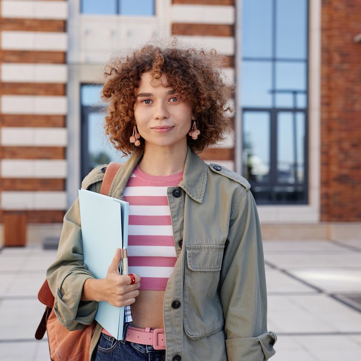 Young woman with curly hair standing in front of a modern building, holding a binder in her hand. She is wearing a green jacket and has a shoulder bag.