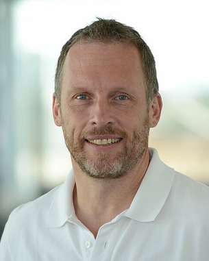 Ein Mann mit kurzem, grauen Haar und Bart trägt ein weißes Polohemd und lächelt freundlich in die Kamera.