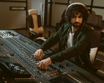 A man with curly hair sits at a mixing console, adjusting the sliders in a recording studio. In the background, other people can be seen.