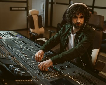 Ein Mann mit lockigem Haar sitzt an einem Mischpult und justiert die Regler in einem Tonstudio. Im Hintergrund sind weitere Personen zu sehen.