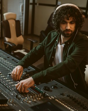 A man with curly hair sits at a mixing console, adjusting the sliders in a recording studio. In the background, other people can be seen.