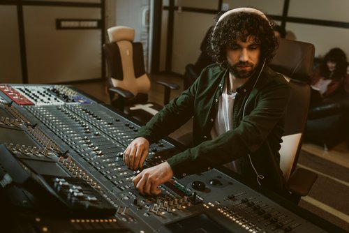 Ein Mann mit lockigem Haar sitzt an einem Mischpult und justiert die Regler in einem Tonstudio. Im Hintergrund sind weitere Personen zu sehen.