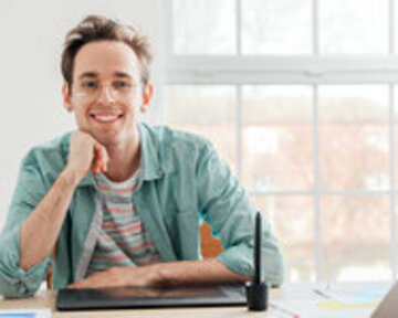 A young man sits at a desk, smiling and resting his chin on his hand. In front of him is a graphic tablet.