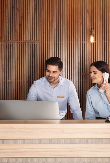 Two employees at a reception desk, one at the computer while the other is on the phone. Wooden paneling and modern lighting in the background.