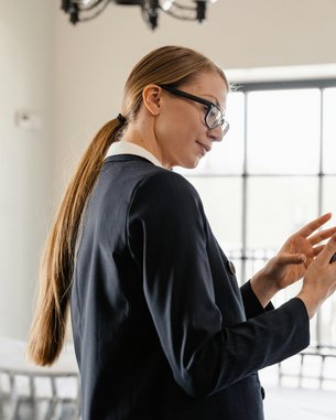 Eine Frau mit langen, glatten Haaren und Brille trägt einen Anzug und schaut auf ihr Smartphone. Sie steht in einem hellen Raum mit großen Fenstern und einem modernen, minimalistischen Design.